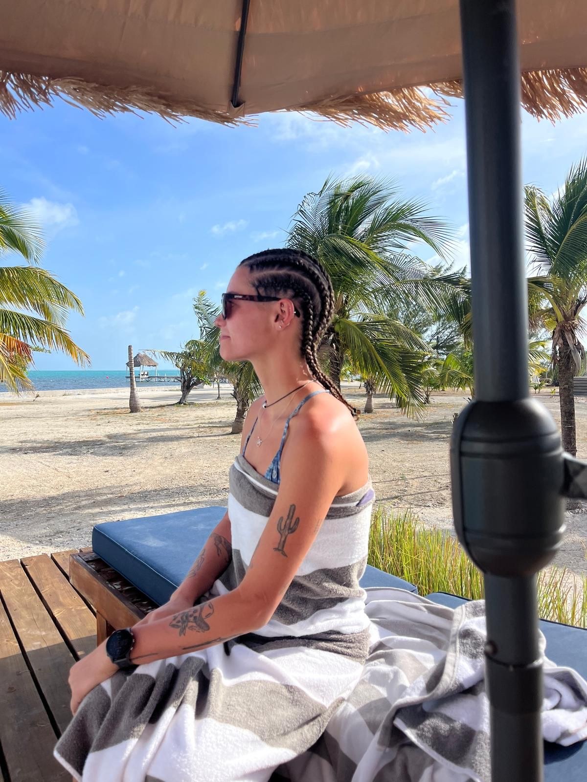 Guest with fresh braids relaxing on a cushioned lounger under a thatched umbrella, with palm trees and the sea behind